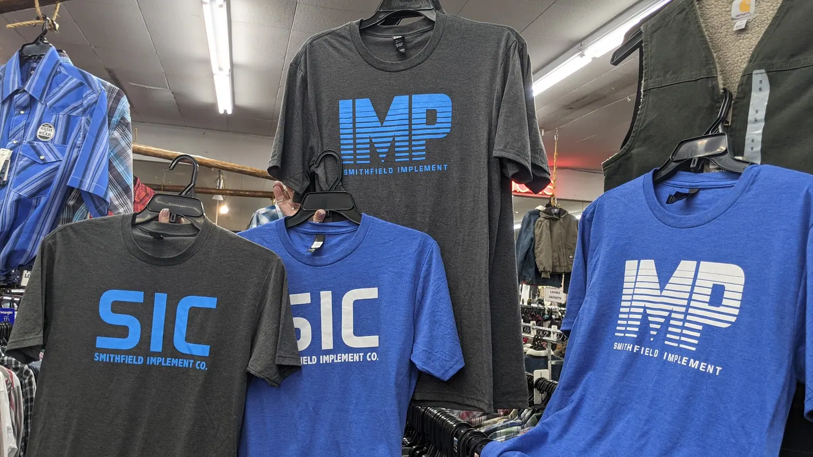 Blue and grey IMP and SIC Smithfield Implement t-shirts hanging on a rack